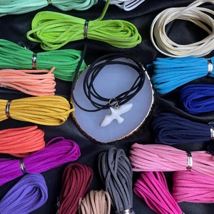 May include: Assortment of colorful suede cords in various colors, including green, blue, orange, yellow, pink, purple, red, and gray. The cords are arranged in a circle around a white ceramic pendant with a bird design.