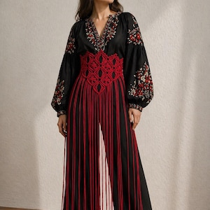 May include: Black jumpsuit with red macrame belt and fringe. The long-sleeved top features floral embroidery around the neckline and sleeves. The wide-leg trousers have long red fringe detailing.