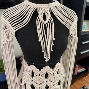 May include: A beige macrame garment featuring a high neckline with radiating cord patterns, a central knotted pendant detail, and long fringe sleeves. The waist section displays a repeating diamond knot pattern with additional hanging fringe.