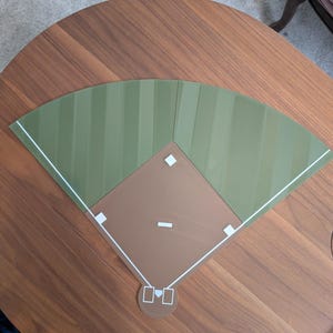 May include: A flat, two-piece baseball field model featuring a brown infield with white base markers and a home plate, connected to a green outfield with subtle striped patterns and white foul lines, forming a complete diamond shape.