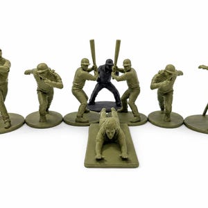 Peut inclure: Un ensemble de sept figurines de baseball monochromes dans diverses poses d'action. Six figurines sont vert olive et une figurine centrale est noire. Chaque figurine est montée sur un socle, représentant des joueurs frappant, courant et glissant.