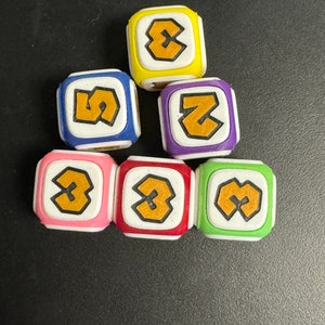 May include: A selection of colourful dice with rounded edges, each showing the number 3 in a bold, orange font. The dice are in various colours, including yellow, blue, purple, pink, red, and green, with white borders. The dice are arranged on a dark surface.