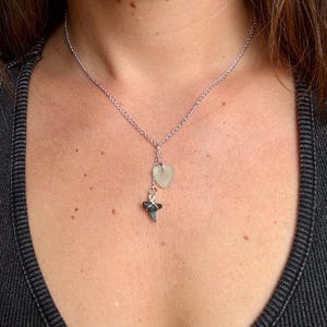 May include: A silver-toned chain necklace featuring two dangling charms: a small, translucent, pale green heart-shaped piece of sea glass and a dark gray, triangular shark tooth wrapped in thin silver-colored wire, suspended from a central link.