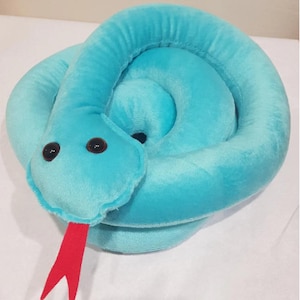 May include: A coiled turquoise plush snake toy. It features large, dark brown eyes and a bright red, forked tongue. The soft toy is designed to resemble a snake.