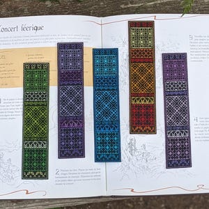 May include: Five colorful bookmarks with intricate geometric patterns. The bookmarks are made of fabric and have a black background with different colored accents. The bookmarks are laid out on a page of a book with the title "Concert féerique" visible.