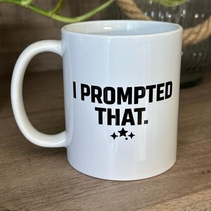 May include: White ceramic mug with a curved handle. The mug features the text "I PROMPTED THAT." in bold black letters, with three black stars below the text. The mug is sitting on a wooden surface.