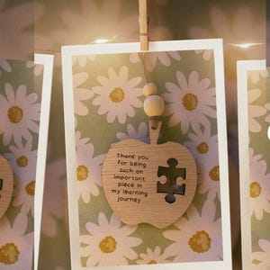 May include: A framed print with a daisy pattern background, held by a wooden clip. A wooden apple-shaped ornament with a puzzle piece cutout and the text "Thank you for being such an important piece in my learning journey" hangs below.