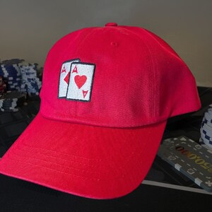 May include: A red baseball cap with an embroidered design of two playing cards, the ace of spades and the ace of hearts.