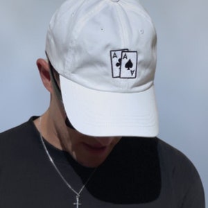Peut inclure: Une casquette de baseball blanche avec un motif brodé noir de deux cartes à jouer, l'as de pique. Le texte "ALL-IN" est brodé sur un t-shirt noir.