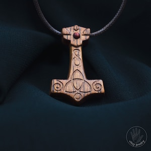 Könnte beinhalten: Eine Holz-Halskette mit einem geschnitzten Mjolnir-Hammer mit einem roten Steineinlege. Der Hammer ist von einem keltischen Knotendesign umgeben.