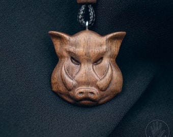 May include: A wooden pendant necklace featuring a carved boar's head. The pendant is a medium brown color, with detailed carving of the boar's tusks, eyes, and snout. The necklace has a black cord and a wooden bead.