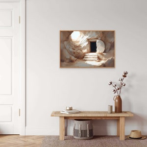 May include: A painting of a stone tomb with a large stone rolled away, revealing a dark opening. The tomb is set against a light background with a bright light shining from the opening. The painting is framed in a light wood frame.