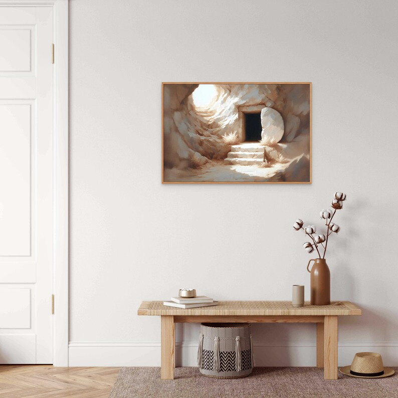 May include: A painting of a stone tomb with a large stone rolled away, revealing a dark opening. The tomb is set against a light background with a bright light shining from the opening. The painting is framed in a light wood frame.