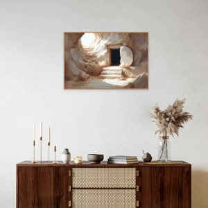 May include: A painting of a stone tomb with a large stone rolled away, revealing a dark opening. The tomb is lit by a bright light coming from the opening. The painting is framed in a light brown frame.
