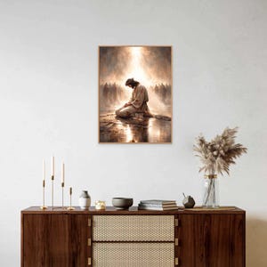May include: A framed print depicts a figure kneeling in prayer, bathed in light, with a crowd in the background. The artwork has a warm color palette and is displayed above a wooden cabinet with decorative items.