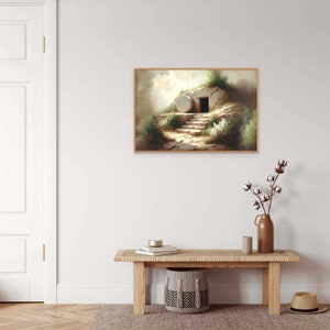 May include: A painting of a stone tomb with a large, round stone rolled away from the entrance. The tomb is set against a backdrop of a rocky hillside with green foliage and a bright, cloudy sky. The painting is framed in a light wood frame.