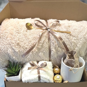 May include: A gift box filled with a cozy white blanket, a white towel with a brown ribbon, a small potted succulent, a white mug with a burlap bag, and a box of gold chocolate candies.