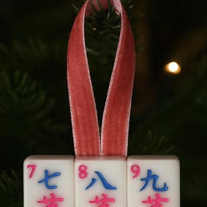 May include: A Christmas ornament made of three white Mahjong tiles, each displaying a number and Chinese characters in blue and pink. The tiles are suspended by a red velvet ribbon. The numbers are 7, 8, and 9.