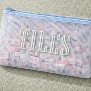 Puede incluir: Una bolsa con cremallera de malla transparente con la palabra "TILES" en letras verde claro. La bolsa est&aacute; llena de peque&ntilde;as baldosas cuadradas en tonos rosa, blanco y azul. La cremallera y el ribete son de color azul claro.