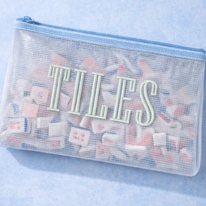 Puede incluir: Una bolsa rectangular semitransparente con cremallera y ribete azules, llena de peque&ntilde;as baldosas de colores. La palabra "TILES" est&aacute; impresa en verde claro en la parte delantera de la bolsa. Las baldosas son rosas, blancas y azules.