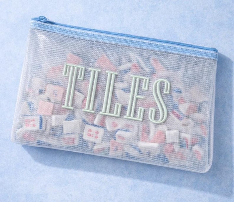Puede incluir: Una bolsa rectangular semitransparente con cremallera y ribete azules, llena de peque&ntilde;as baldosas de colores. La palabra "TILES" est&aacute; impresa en verde claro en la parte delantera de la bolsa. Las baldosas son rosas, blancas y azules.