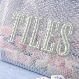 Puede incluir: Una bolsa de malla con la palabra "TILES" bordada en verde claro. La bolsa est&aacute; llena de peque&ntilde;as baldosas de colores. Tres baldosas blancas con n&uacute;meros azules est&aacute;n delante de la bolsa.