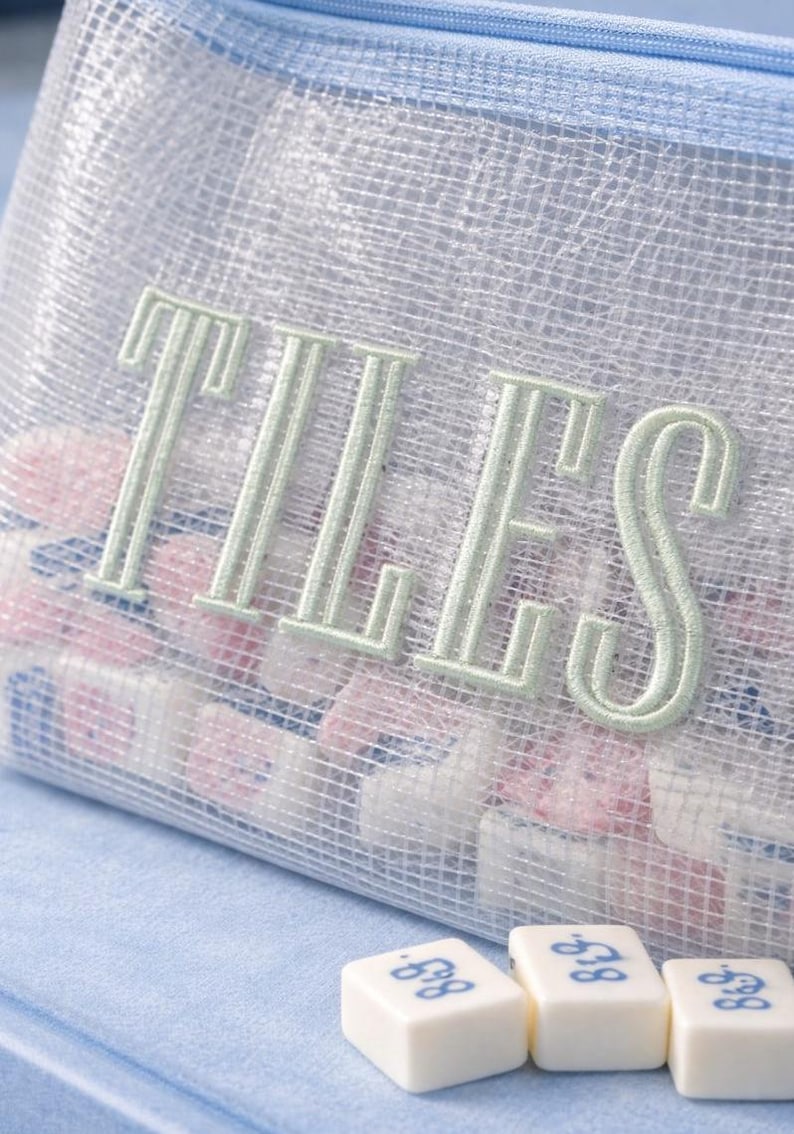 Puede incluir: Una bolsa de malla con la palabra "TILES" bordada en verde claro. La bolsa est&aacute; llena de peque&ntilde;as baldosas de colores. Tres baldosas blancas con n&uacute;meros azules est&aacute;n delante de la bolsa.