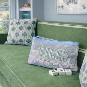 May include: A green sofa with a patterned cushion and a grey pouch embroidered with the word "TILES" in green. Three white game tiles are in front of the pouch. The pouch has a blue zip.