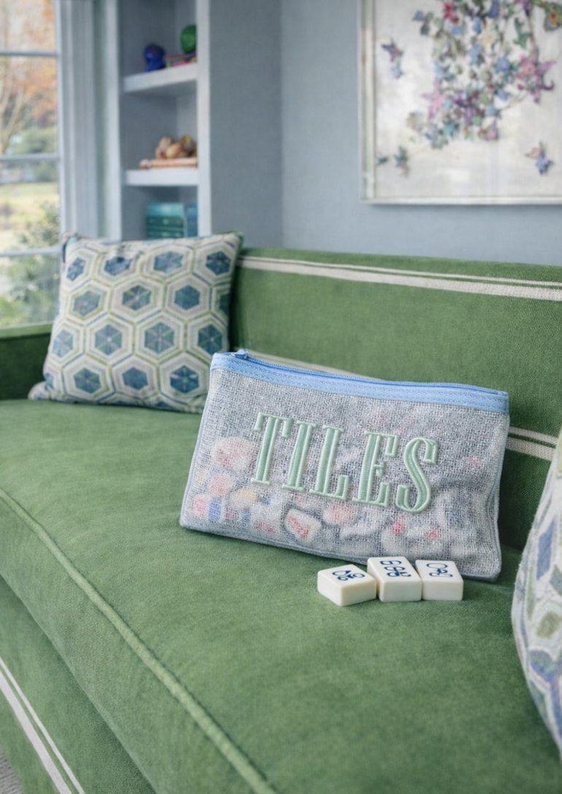 Puede incluir: Un sof&aacute; verde con un coj&iacute;n estampado y una bolsa gris bordada con la palabra "TILES" en verde. Tres fichas de juego blancas est&aacute;n frente a la bolsa. La bolsa tiene una cremallera azul.