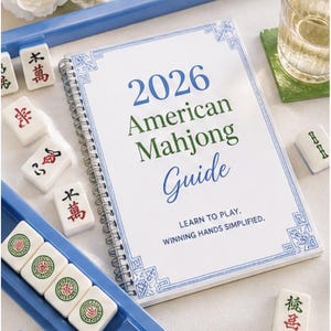 May include: A spiral-bound "2026 American Mahjong Guide" book with blue and green text, surrounded by white Mahjong tiles and a blue tray. The text "Learn to Play. Winning Hands Simplified" is also visible. A glass of liquid sits on a green coaster.