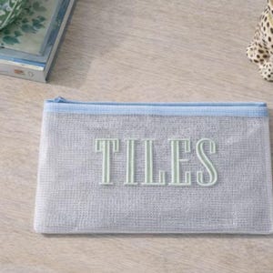 Puede incluir: Una bolsa de malla gris claro con cremallera y ribete azul claro. La palabra "TILES" est&aacute; bordada en color verde claro en la parte delantera. Un portavelas decorativo con estampado de leopardo est&aacute; en el fondo.