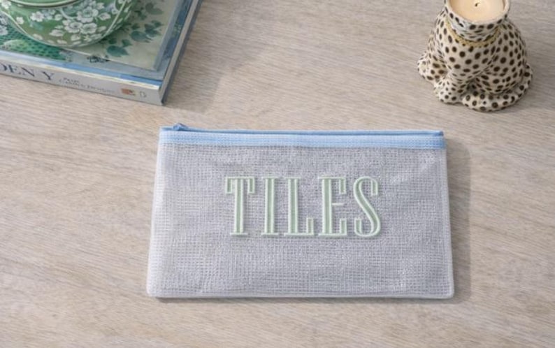 Puede incluir: Una bolsa de malla gris claro con cremallera y ribete azul claro. La palabra "TILES" est&aacute; bordada en color verde claro en la parte delantera. Un portavelas decorativo con estampado de leopardo est&aacute; en el fondo.