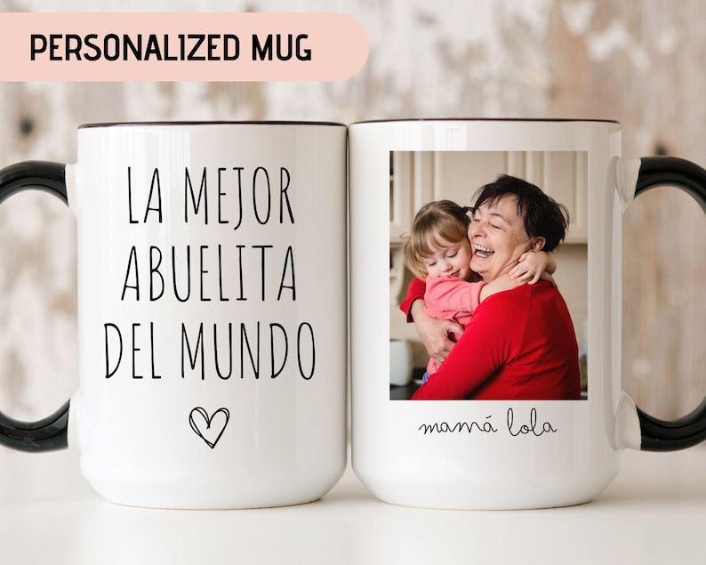 May include: White ceramic mug with a black handle. The mug has a photo of a grandmother and granddaughter hugging on one side and the text "LA MEJOR ABUELITA DEL MUNDO" with a heart on the other side. The text below the photo reads "mam&aacute; lola".