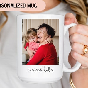 May include: White ceramic mug with a photo of a grandmother and granddaughter hugging. The text "mam&aacute; lola" is printed on the mug.