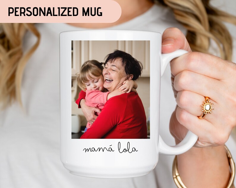 May include: White ceramic mug with a photo of a grandmother and granddaughter hugging. The text "mam&aacute; lola" is printed on the mug.