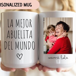 May include: White ceramic mug with a black inscription that reads "LA MEJOR ABUELITA DEL MUNDO" with a small heart at the bottom. The mug has a photo of a grandmother and granddaughter hugging, with the text "mam&aacute; lola" below the photo.
