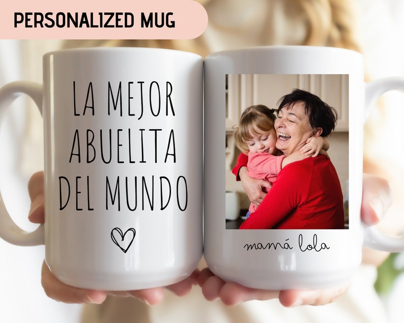 May include: White ceramic mug with a black inscription that reads "LA MEJOR ABUELITA DEL MUNDO" with a small heart at the bottom. The mug has a photo of a grandmother and granddaughter hugging, with the text "mam&aacute; lola" below the photo.