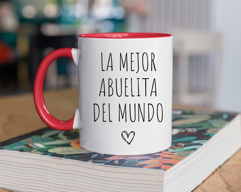 May include: White ceramic mug with a red rim and handle. The mug has black text that reads "LA MEJOR ABUELITA DEL MUNDO" with a small heart symbol.