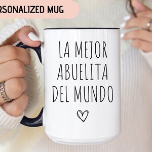 May include: White ceramic mug with a black rim and handle. The mug has black text that reads "LA MEJOR ABUELITA DEL MUNDO" with a small heart symbol below the text.