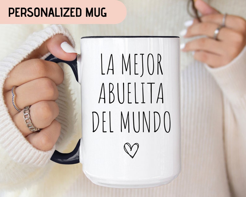 May include: White ceramic mug with a black rim and handle. The mug has black text that reads "LA MEJOR ABUELITA DEL MUNDO" with a small heart symbol below the text.