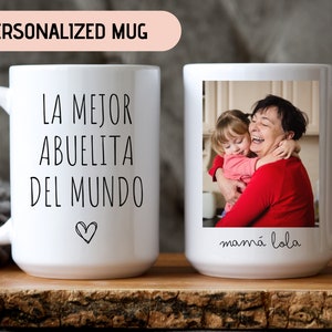 May include: White ceramic mug with a black inscription that reads "La mejor abuelita del mundo" with a heart symbol. The mug has a photo of a grandmother and granddaughter hugging, with the text "mam&aacute; lola" below the photo.