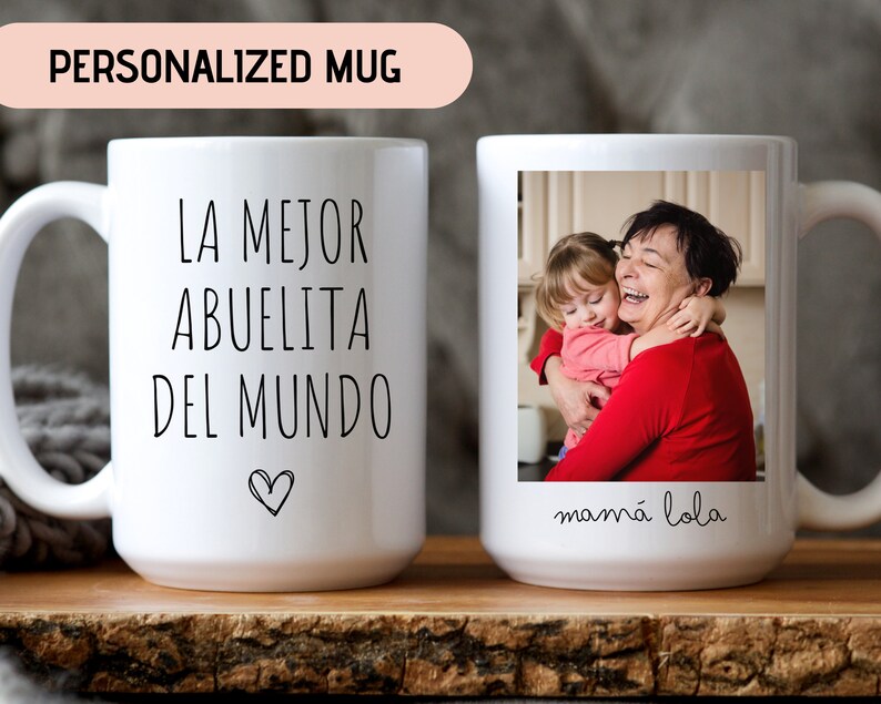 May include: White ceramic mug with a black inscription that reads "La mejor abuelita del mundo" with a heart symbol. The mug has a photo of a grandmother and granddaughter hugging, with the text "mam&aacute; lola" below the photo.