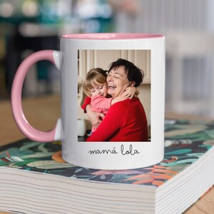 May include: White ceramic mug with a pink rim and handle. The mug features a photo of a grandmother and granddaughter hugging, with the text "mam&aacute; lola" printed below the photo.