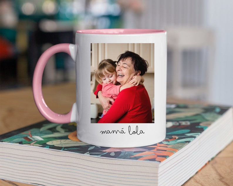 May include: White ceramic mug with a pink rim and handle. The mug features a photo of a grandmother and granddaughter hugging, with the text "mam&aacute; lola" printed below the photo.