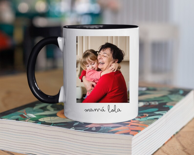 May include: White ceramic mug with a black handle and a black rim. The mug features a photo of a grandmother and granddaughter hugging, with the text "mamma lola" written below the photo.