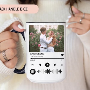 May include: White ceramic mug with a black handle. The mug features a photo of a couple in a garden setting. The text "SARA'S SONG" is printed on the mug, along with the words "You're my soulmate". The mug also has a Spotify-like music player interface with a play button and a progress bar.