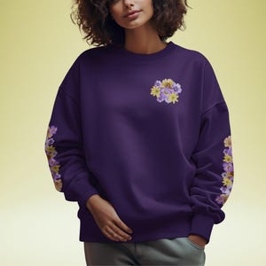 May include: A deep purple sweatshirt featuring floral embroidery on the left sleeve and chest. The flowers are yellow, purple, and white. The sweatshirt has a crew neck and long sleeves.