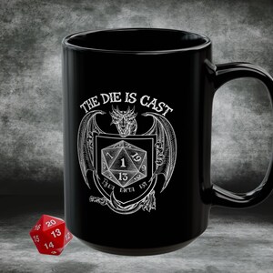 May include: Black ceramic mug with a white graphic that reads "THE DIE IS CAST" and features a dragon, a shield, and a 20-sided die. A red 20-sided die is next to the mug. The mug is designed for tabletop role-playing games.