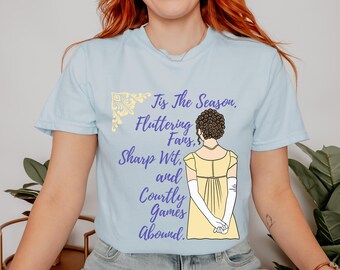Könnte beinhalten: Hellblaues T-Shirt mit einem lila und gelben Design. Das Design zeigt eine Frau in einem gelben Kleid mit dem Text "Tis the Season, Fluttering Fans, Sharp Wit, and Courtly Games Abound."