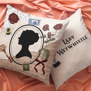 May include: Two decorative throw pillows with a vintage aesthetic. One pillow features a black silhouette of a woman in an oval frame, a key, a bee, and floral accents. The other pillow displays the text "LADY WETWHISTLE".